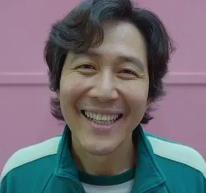 Um homem sorridente de agasalho verde em destaque. Ao fundo, figura mascarada de rosa em ambiente rosa. Clima de alegria, mas ameaçador.