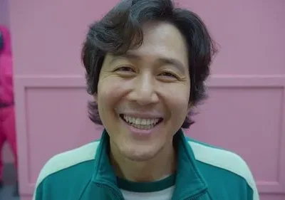 Um homem sorridente de agasalho verde em destaque. Ao fundo, figura mascarada de rosa em ambiente rosa. Clima de alegria, mas ameaçador.