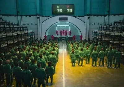 Multidão de pessoas em uniformes verdes olha para figuras mascaradas de vermelho em salão com beliches. Placar eletrônico indica jogo tenso.