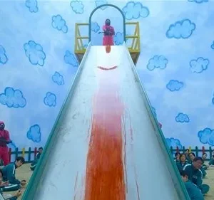 Pessoas em macacões verdes sentadas num quarto azul com nuvens. Guardas mascarados em vermelho, com armas. Um escorrega com mancha de sangue. Clima tenso e sinistro.