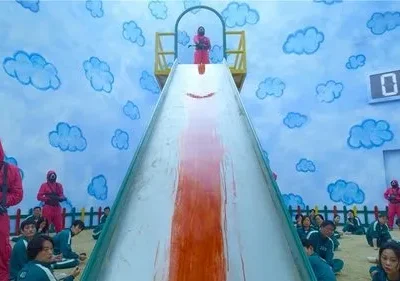 Pessoas em macacões verdes sentadas num quarto azul com nuvens. Guardas mascarados em vermelho, com armas. Um escorrega com mancha de sangue. Clima tenso e sinistro.