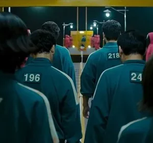 Jogadores de uniforme numerado verde-azulado caminham em fila por um corredor sombrio. Guardas de capuz vermelho observam.