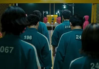 Jogadores de uniforme numerado verde-azulado caminham em fila por um corredor sombrio. Guardas de capuz vermelho observam.