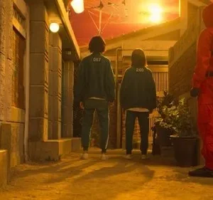 Duas figuras de uniforme verde e uma de macacão vermelho em um beco. Céu laranja vibrante. Atmosfera tensa.