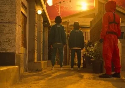 Duas figuras de uniforme verde e uma de macacão vermelho em um beco. Céu laranja vibrante. Atmosfera tensa.