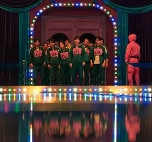 Pessoas em uniformes verdes numerados num palco vibrante. Um guarda de rosa os observa. Cenário teatral com luzes coloridas e reflexos.