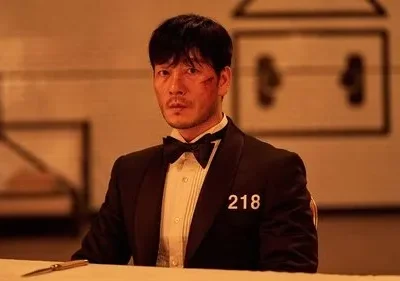 Homem sério de smoking com nº 218 e ferida no rosto. Sentado à mesa em ambiente laranja e tenso com detalhes borrados.