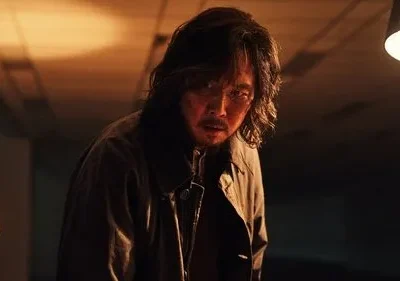 Homem de cabelo bagunçado e casaco, rosto sujo e tenso, em ambiente escuro com iluminação dramática e contrastante.