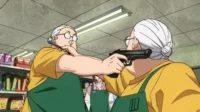 Duas idosas de avental em um mercado. Uma aponta uma pistola para a cabeça da outra. Cena tensa e inesperada.