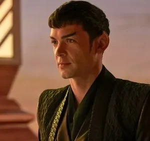 Homem de orelhas pontudas e cabelo tigela, em manto escuro. Ele tem um olhar sério. Fundo suave com luz dourada e detalhe arquitetônico.