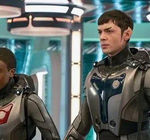 Dois tripulantes em trajes futuristas em uma nave espacial. Um homem com orelhas pontudas e um jovem negro olham atentos.