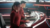 Mulher e homem em uniformes vermelhos operam consoles futuristas iluminados em vermelho, em uma cena de ficção científica.