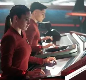 Mulher e homem em uniformes vermelhos operam consoles futuristas iluminados em vermelho, em uma cena de ficção científica.