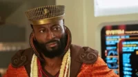 Homem negro barbudo com chapéu dourado ornado, trajes vibrantes e colares, em ambiente futurista. Olhar sério.