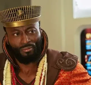 Homem negro barbudo com chapéu dourado ornado, trajes vibrantes e colares, em ambiente futurista. Olhar sério.