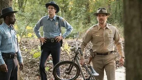 Três homens uniformizados de xerife em um bosque. Um carrega uma bicicleta. Aparência pensativa, clima de investigação.