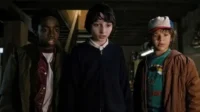 Três meninos, um de boné, olham tensos em um ambiente escuro e misterioso, iluminados por uma luz fraca.