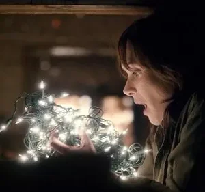 Mulher em perfil, surpresa, segura luzes de Natal brilhantes que iluminam seu rosto em ambiente escuro.