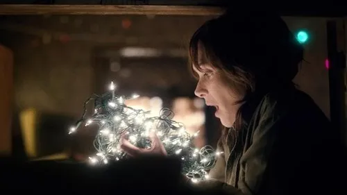Mulher em perfil, surpresa, segura luzes de Natal brilhantes que iluminam seu rosto em ambiente escuro.