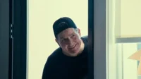 Homem sorridente de boné escuro espreita por trás de um batente de porta, em um ambiente claro.