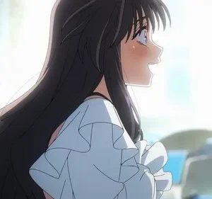 Jovem anime de cabelos escuros e longos, sorrindo e corando, com olhar de surpresa. Fundo interior claro e desfocado, com forte luz.