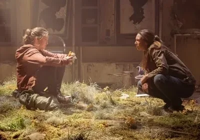 Duas mulheres em um ambiente arruinado com vegetação. Uma sentada, outra agachada. Clima desolado, de interação íntima.