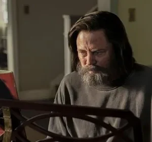 Homem barbudo de cabelo comprido e expressão séria olha para uma partitura musical. Clima introspectivo em um ambiente interno.