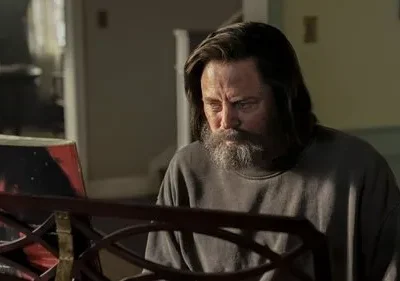 Homem barbudo de cabelo comprido e expressão séria olha para uma partitura musical. Clima introspectivo em um ambiente interno.