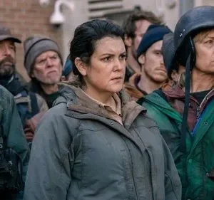 Multidão de sobreviventes sérios, vestindo roupas rústicas e táticas. Um homem com colete, duas mulheres em destaque. Clima tenso e vigilante.