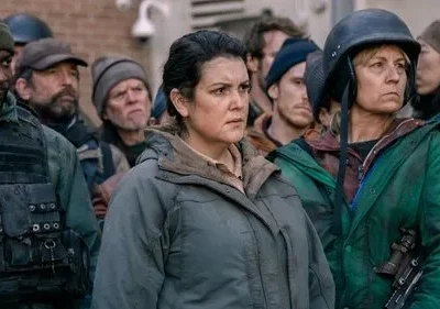 Multidão de sobreviventes sérios, vestindo roupas rústicas e táticas. Um homem com colete, duas mulheres em destaque. Clima tenso e vigilante.