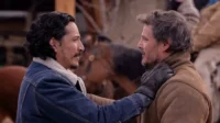 Dois homens em casacos, um de jeans e outro marrom, conversam de perto. Um segura o ombro do outro em fundo rural.