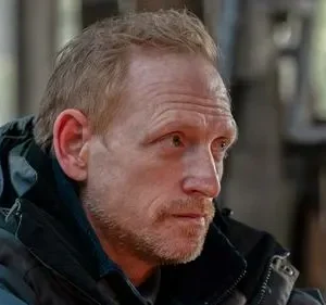 Close-up de homem ruivo barbudo com jaqueta escura, olhando para o lado com expressão séria. Fundo desfocado de estrutura interna.