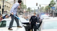 Homem de pijama com bastão sobre carro. Policiais e multidão em rua caótica. Clima cômico de perseguição urbana.