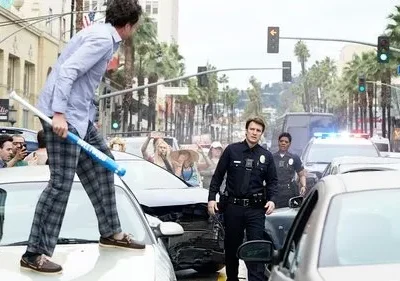 Homem de pijama com bastão sobre carro. Policiais e multidão em rua caótica. Clima cômico de perseguição urbana.