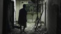 Homem em camisola caminha em corredor escuro e destruído. Portas com DON'T DEAD OPEN INSIDE grafitado. Clima sombrio.