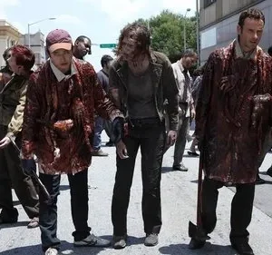 Homens e uma mulher camuflados com sangue e entranhas andam por uma rua com muitos zumbis ao redor. Clima apocalíptico.