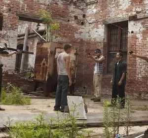 Confronto tenso em pátio abandonado. Dois homens com besta e rifle contra um grupo com pistolas, outro no centro. Ruínas ao fundo.