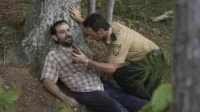 Um xerife agachado ajuda um homem ferido encostado numa árvore na floresta. O clima é de urgência e preocupação.