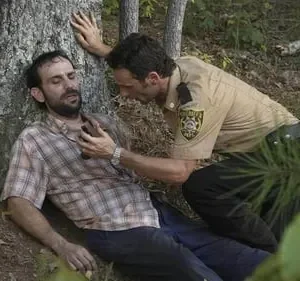 Um xerife agachado ajuda um homem ferido encostado numa árvore na floresta. O clima é de urgência e preocupação.