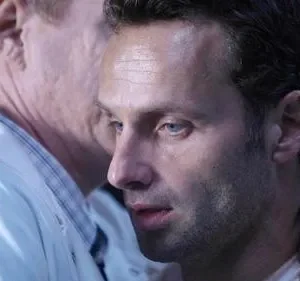 Close de dois homens. Um de camisa branca e outro com barba e olhos azuis, que segura o ombro do primeiro, com semblante aflito em cenário escuro.