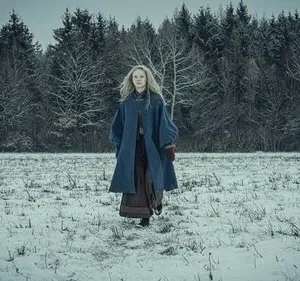 Jovem mulher de cabelos claros em casaco azul, caminhando por um campo nevado com floresta densa e sombria. Clima invernal e misterioso.