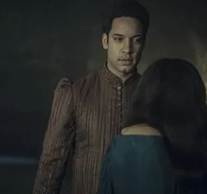 Perto: homem sério em túnica marrom olha mulher de costas em vestido azul. Fundo escuro, uma luz fraca. Clima sombrio.