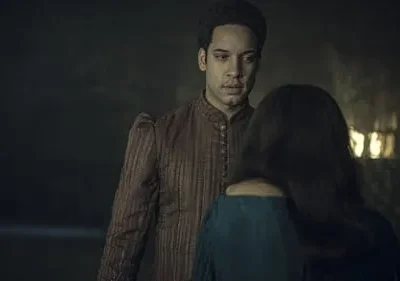 Perto: homem sério em túnica marrom olha mulher de costas em vestido azul. Fundo escuro, uma luz fraca. Clima sombrio.