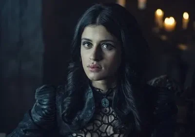 Mulher de cabelos escuros e olhar intenso, vestindo roupa preta elaborada, em ambiente escuro com luzes no fundo. Clima misterioso.