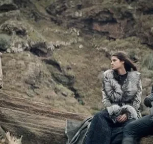 Cena com um homem barbudo, uma mulher de casaco de pele e um homem de cabelos brancos sentados em rochas, paisagem montanhosa.