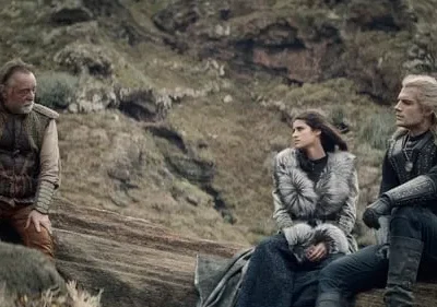 Cena com um homem barbudo, uma mulher de casaco de pele e um homem de cabelos brancos sentados em rochas, paisagem montanhosa.