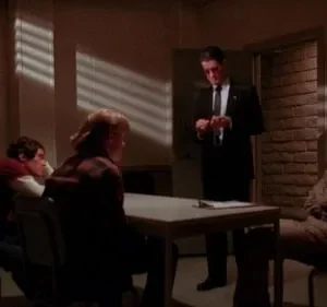 Quatro homens em uma sala escura. Um homem de terno em pé, três sentados. Luz em persianas cria listras. Clima tenso.