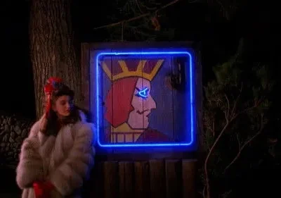 Mulher em casaco de pele branco e luvas vermelhas ao lado de um letreiro neon azul de um rei. Cena noturna misteriosa.