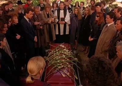 Funeral ao ar livre: Pessoas de luto cercam um caixão vermelho com flores. Um padre lidera, o ambiente é de tristeza profunda.