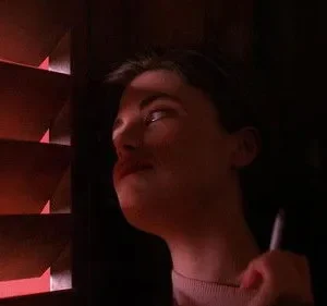 Jovem mulher em quarto escuro, rosto parcialmente iluminado por luz vermelha de persianas. Segura um cigarro, olhar pensativo. Clima de mistério.
